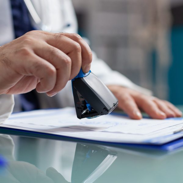 Doctor holding medical stamp to put on prescription paper at health care exam. Hand of physician putting seal on checkup report, giving medicine to patient at consultation. Close up